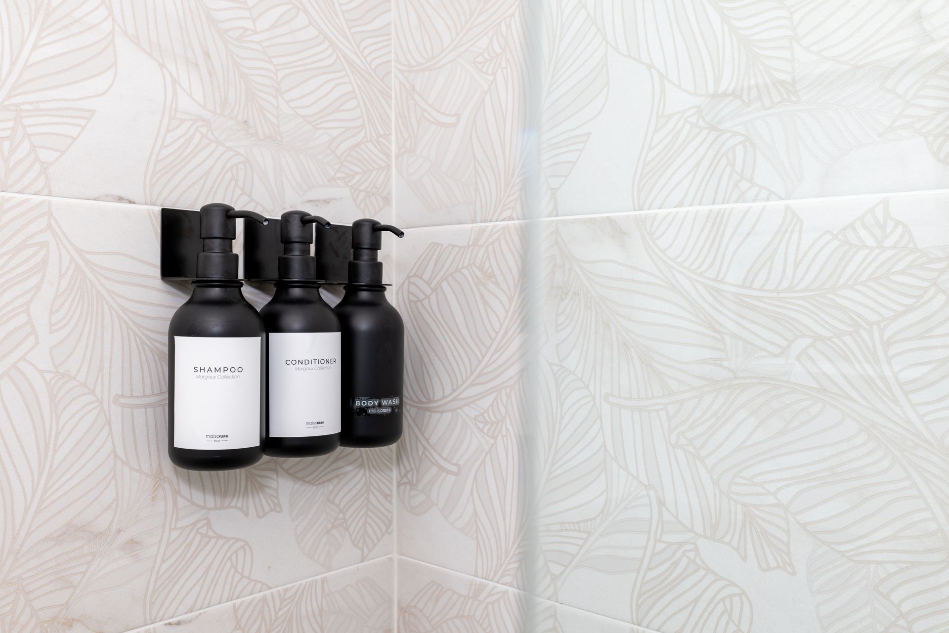 Three black bottles in a shower, labeled with white text, on patterned beige tiles.