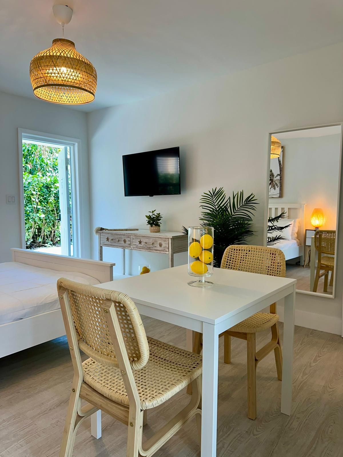 Bright, white-walled room with dining table and chairs.  A TV hangs on the wall. A bedroom is visible through a mirror.