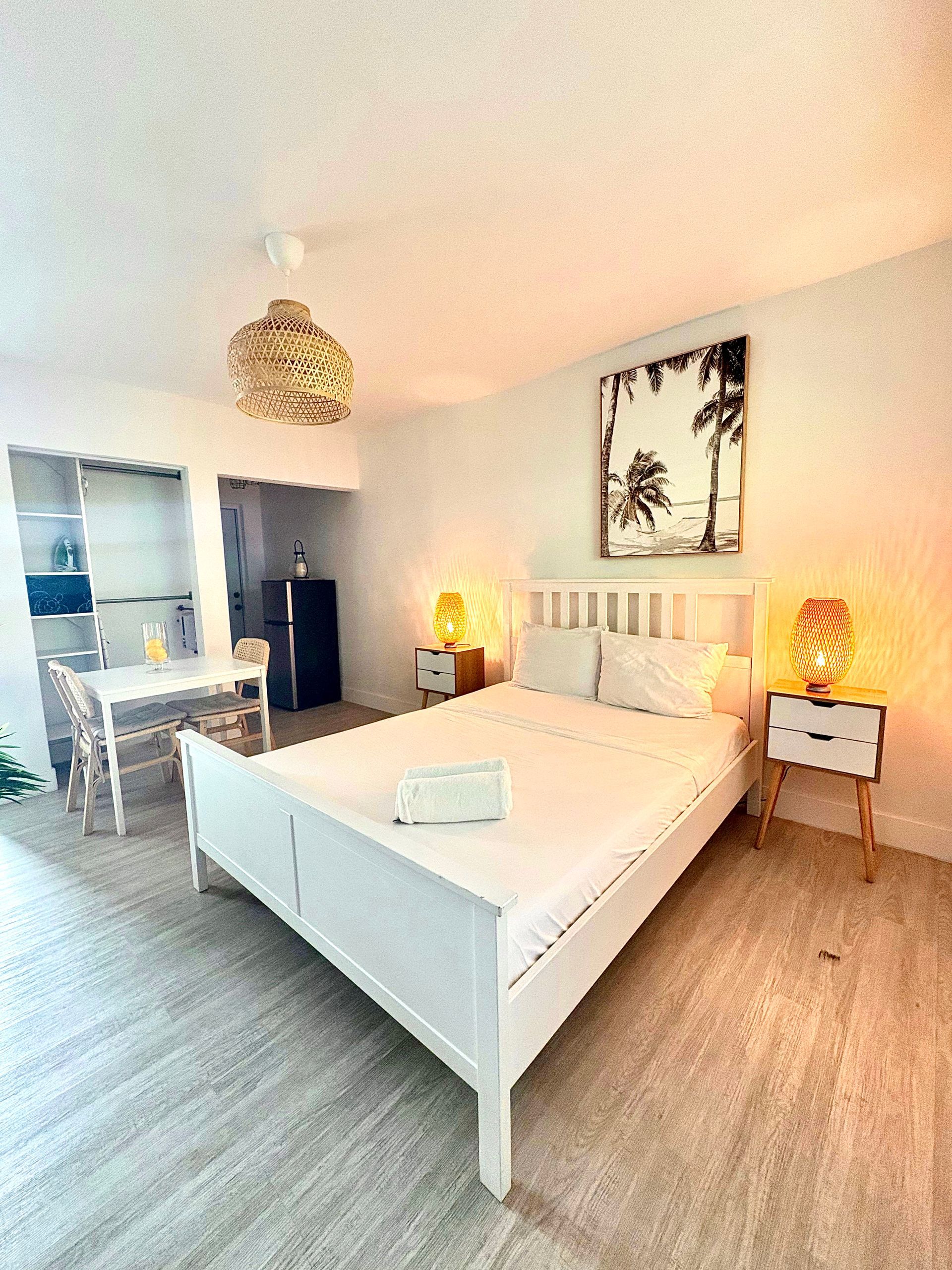 White bedroom with bed, nightstands, dining area, and beach art; lit by warm lamps.