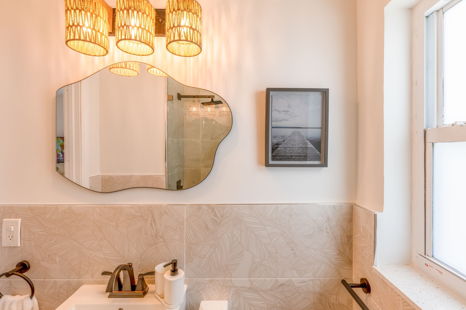 Bathroom with a wavy mirror, art print, and tan-colored tiles and walls. Includes a window and light fixtures.