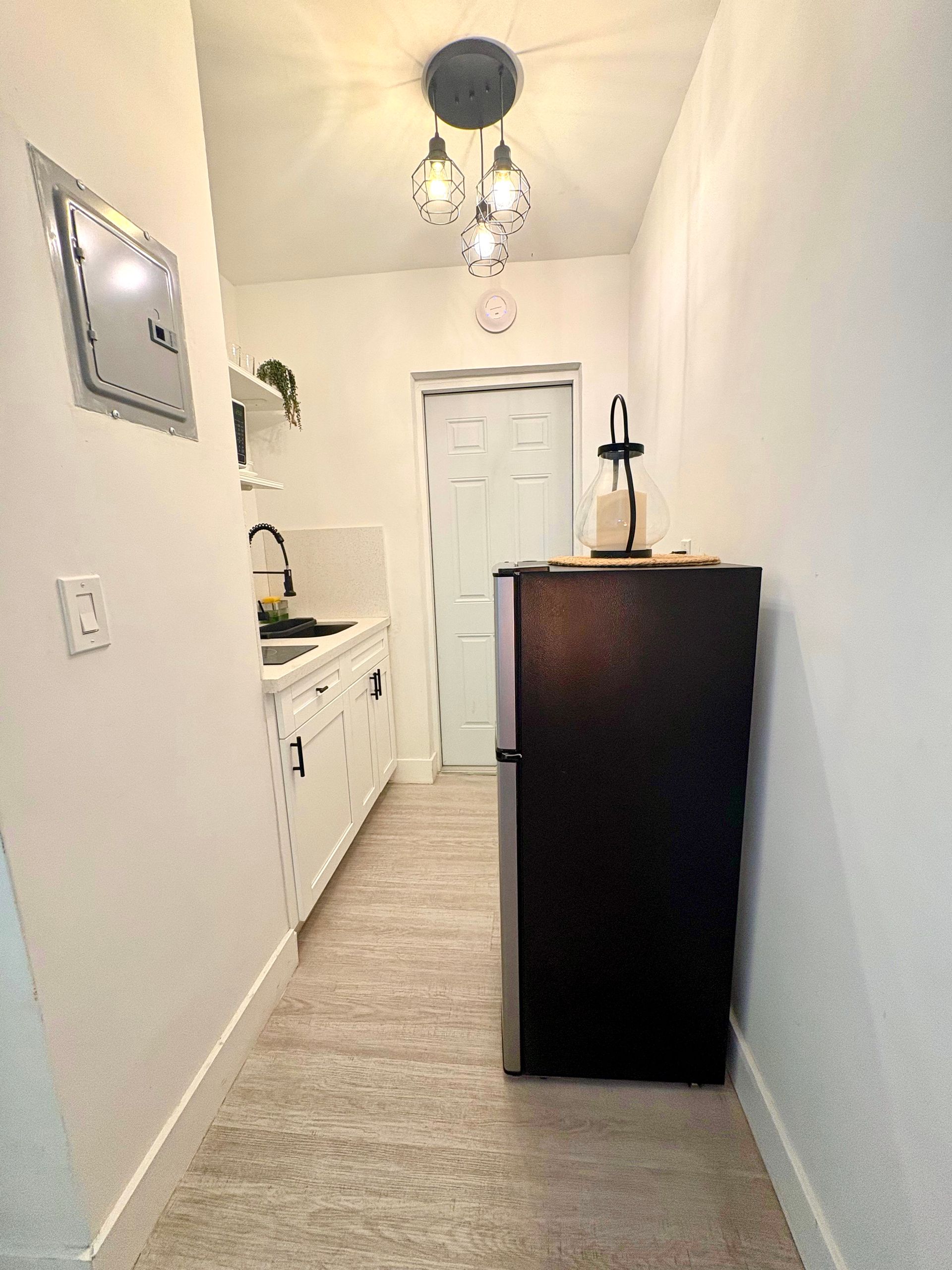 Narrow hallway with kitchenette on the left, a dark refrigerator, and a door at the end.