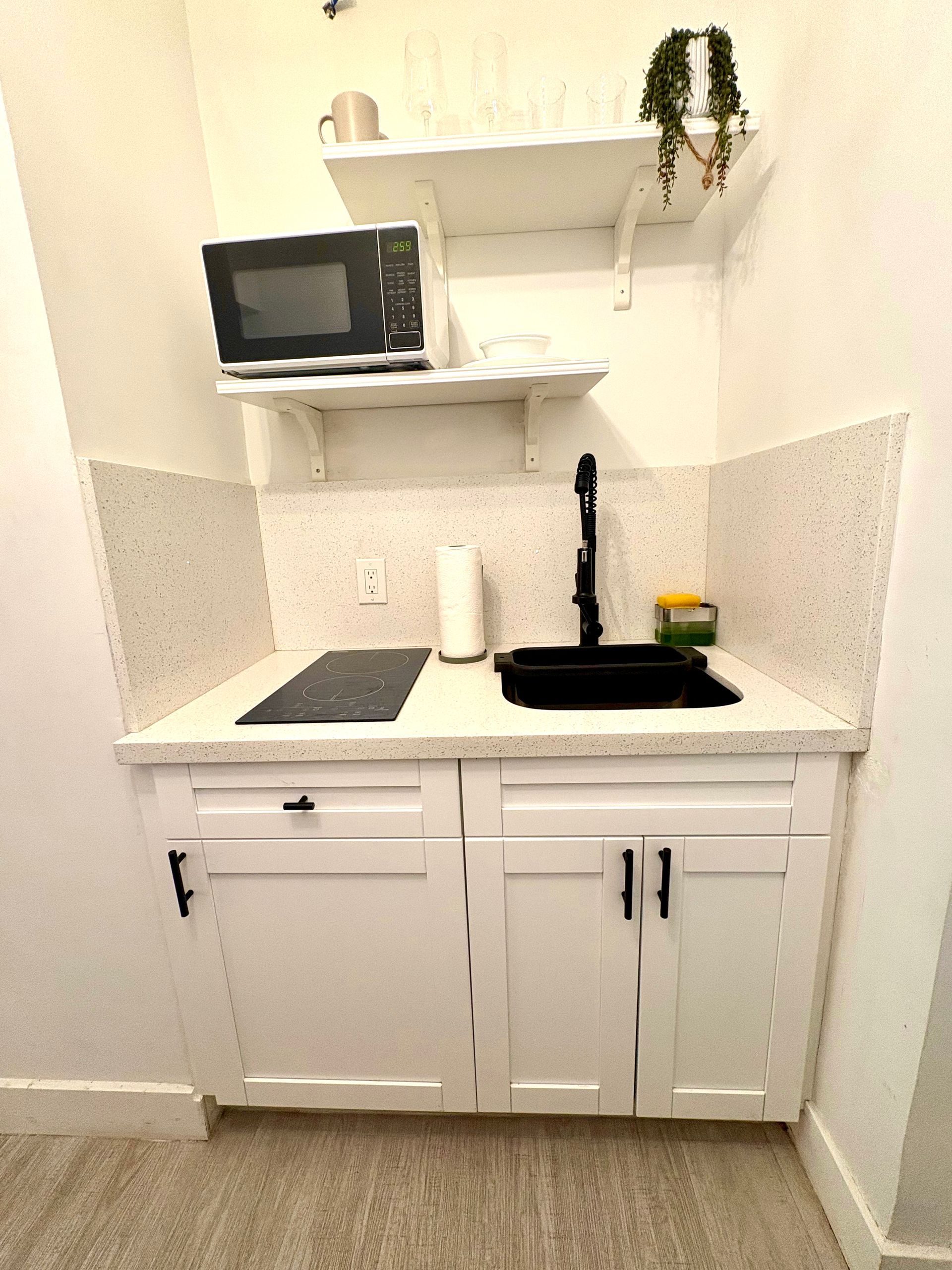 Small white kitchenette with a black sink, microwave, and cooktop. Cabinets and a shelf are present.