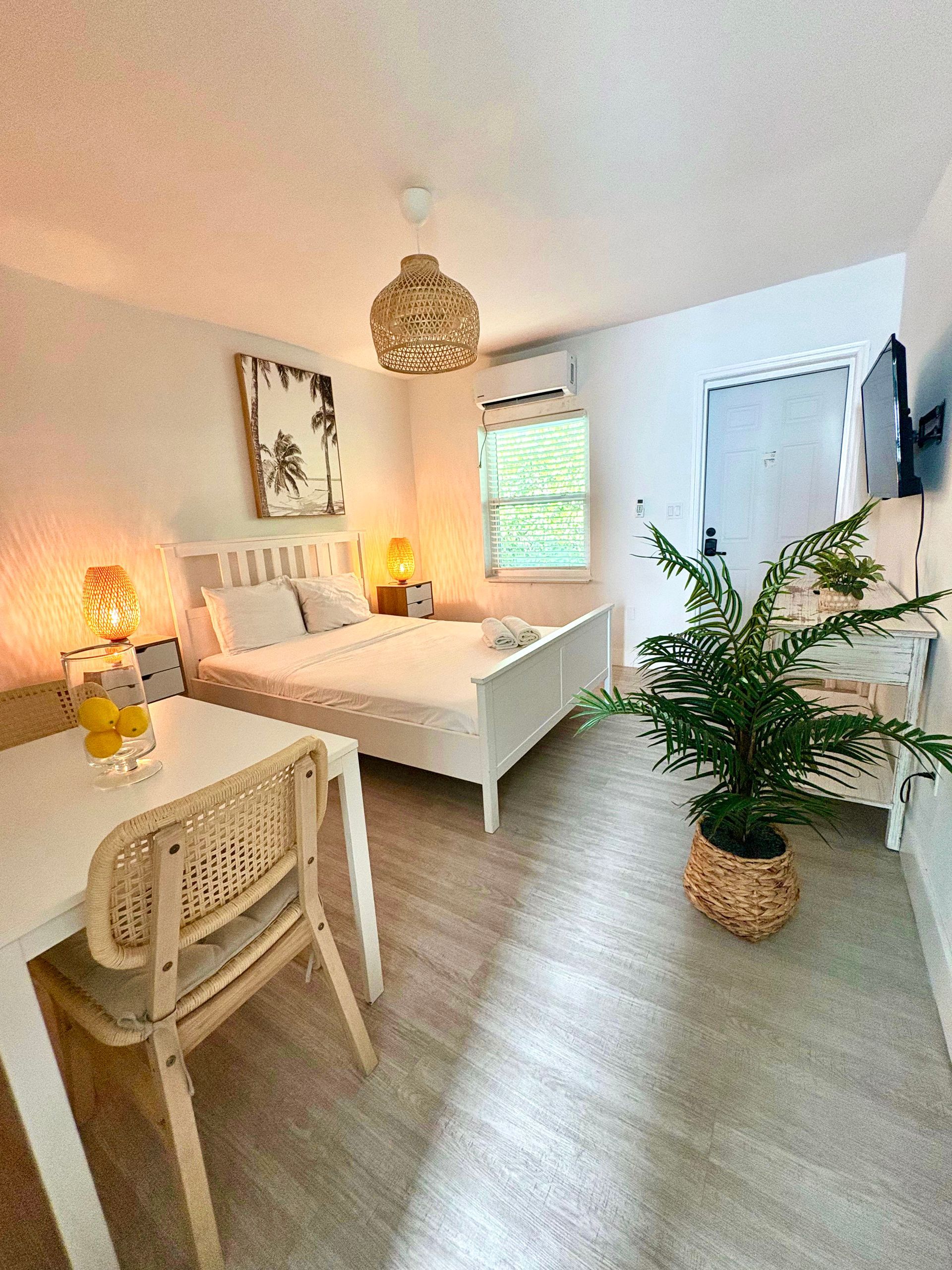 Cozy white studio apartment with bed, table, chair, and decorative lighting, featuring a potted plant.