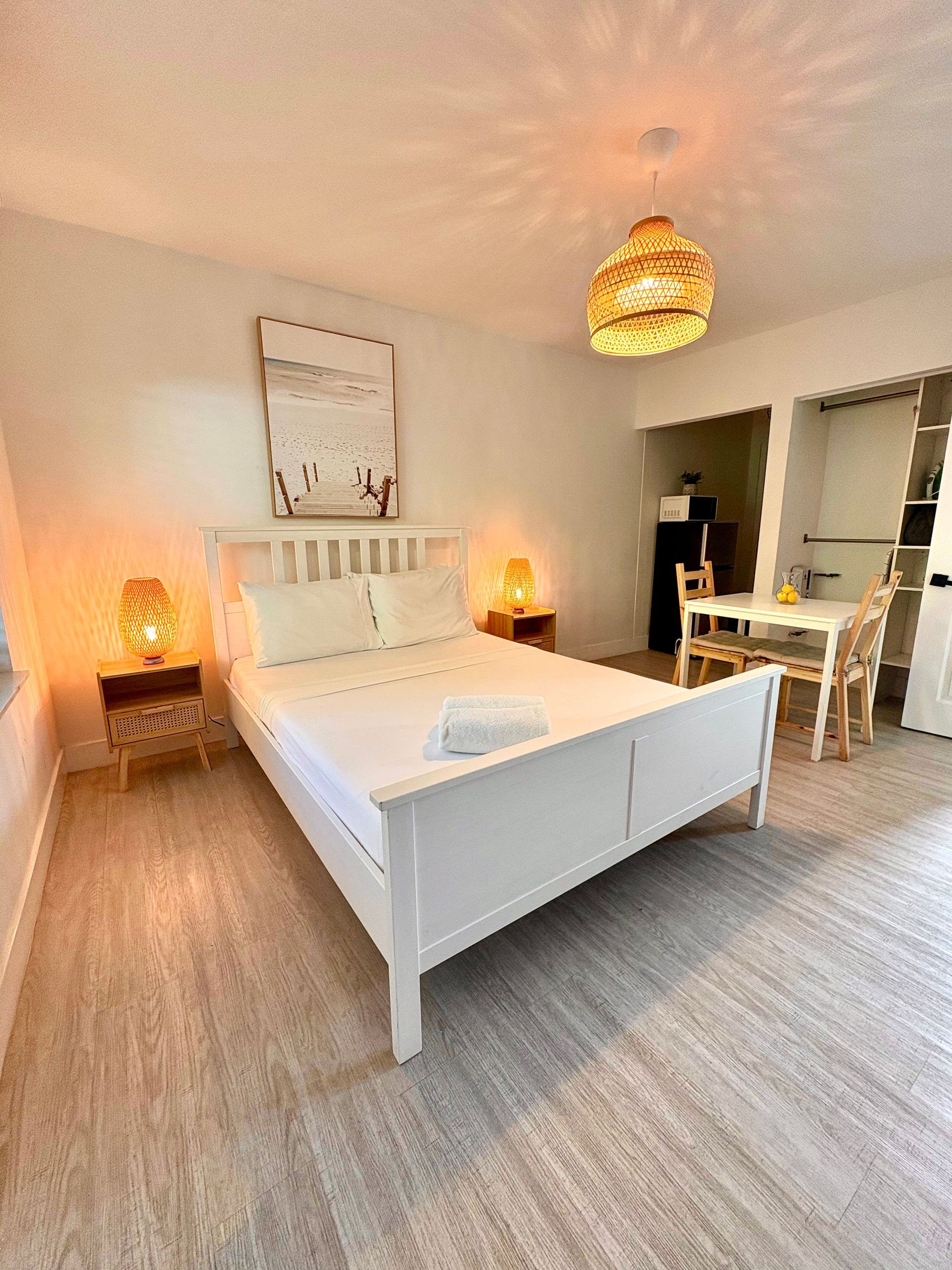 White bedroom with bed, nightstands, artwork, and dining area; warm lighting.