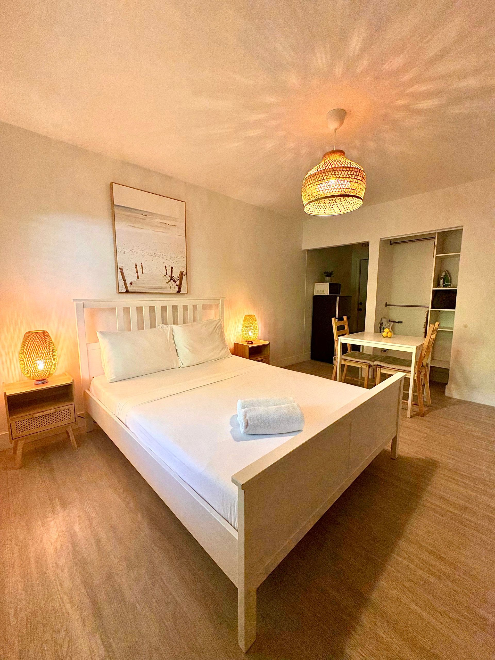 Bedroom with white bed, bedside tables, and a small kitchen area. Warm lighting.