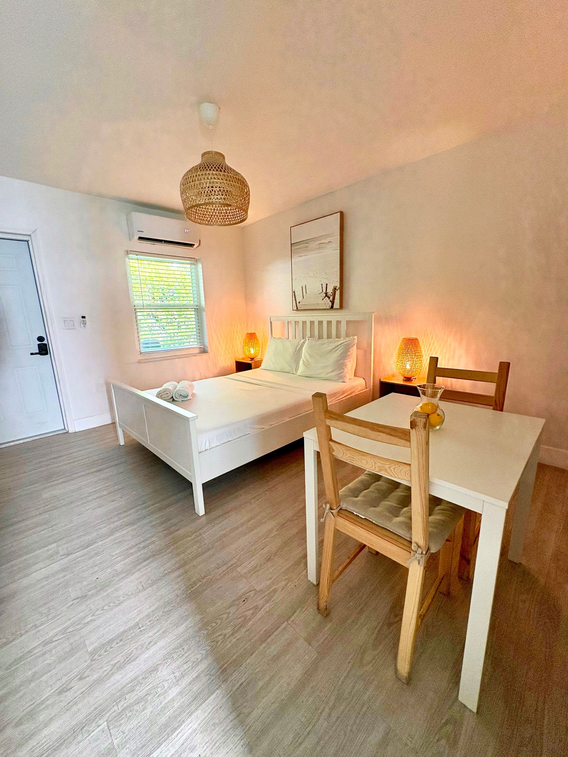 Cozy bedroom with white bed, wooden table, and two chairs. A woven light fixture hangs overhead.