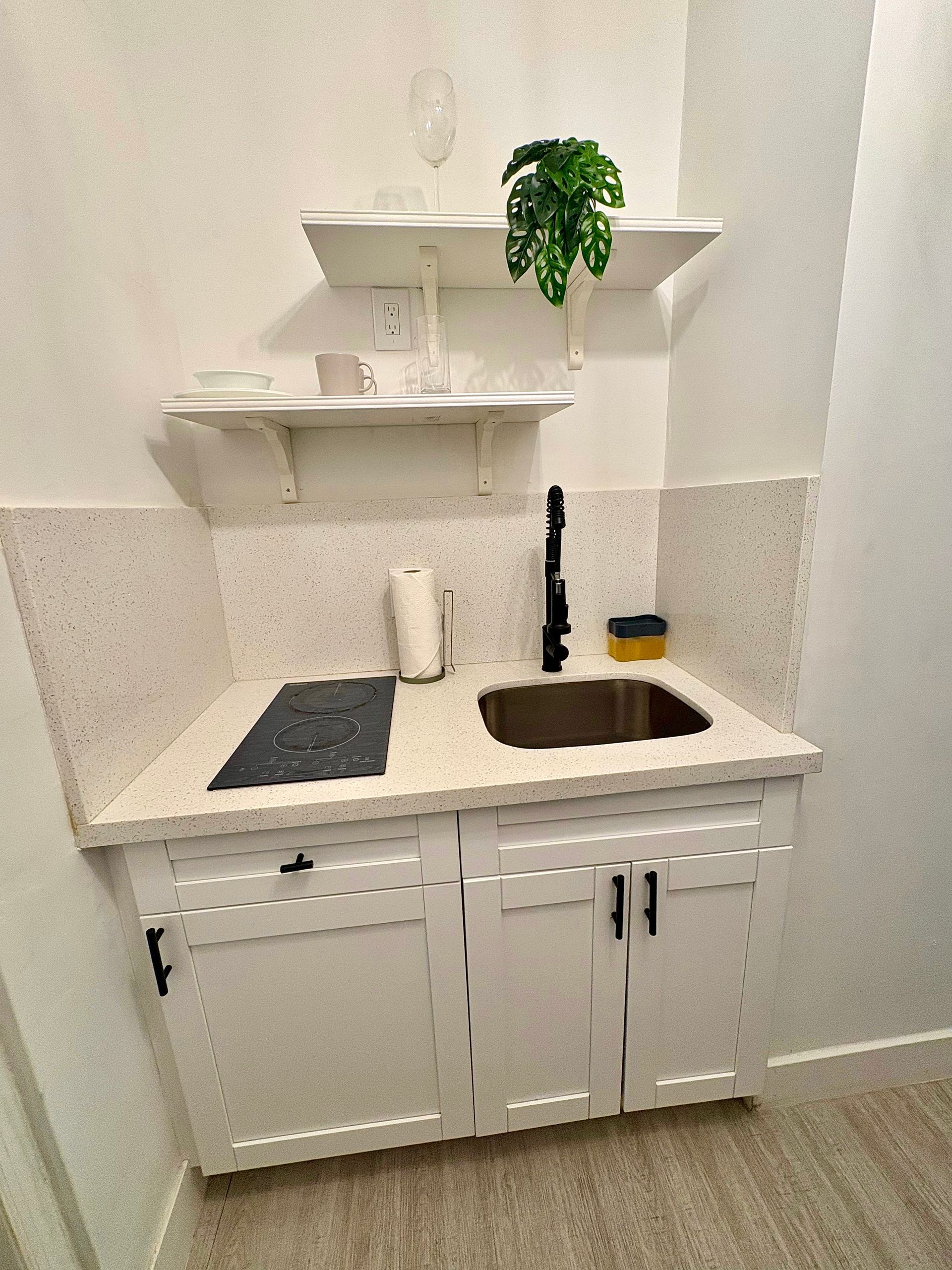 Small white kitchenette with cabinets, countertop, sink, cooktop, and shelves.