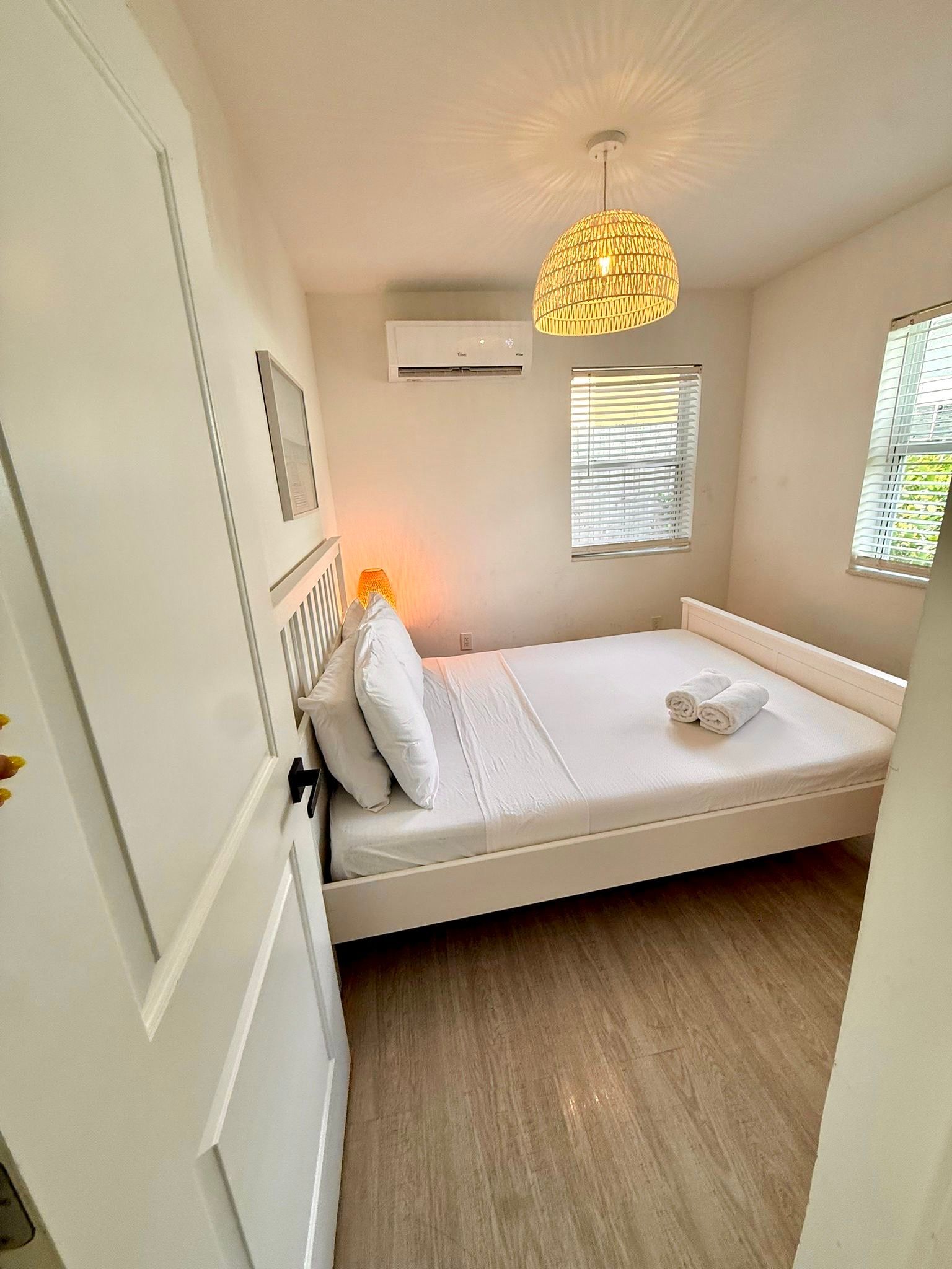 Bright bedroom with a white bed, walls, and door. A decorative light and AC unit are also visible.