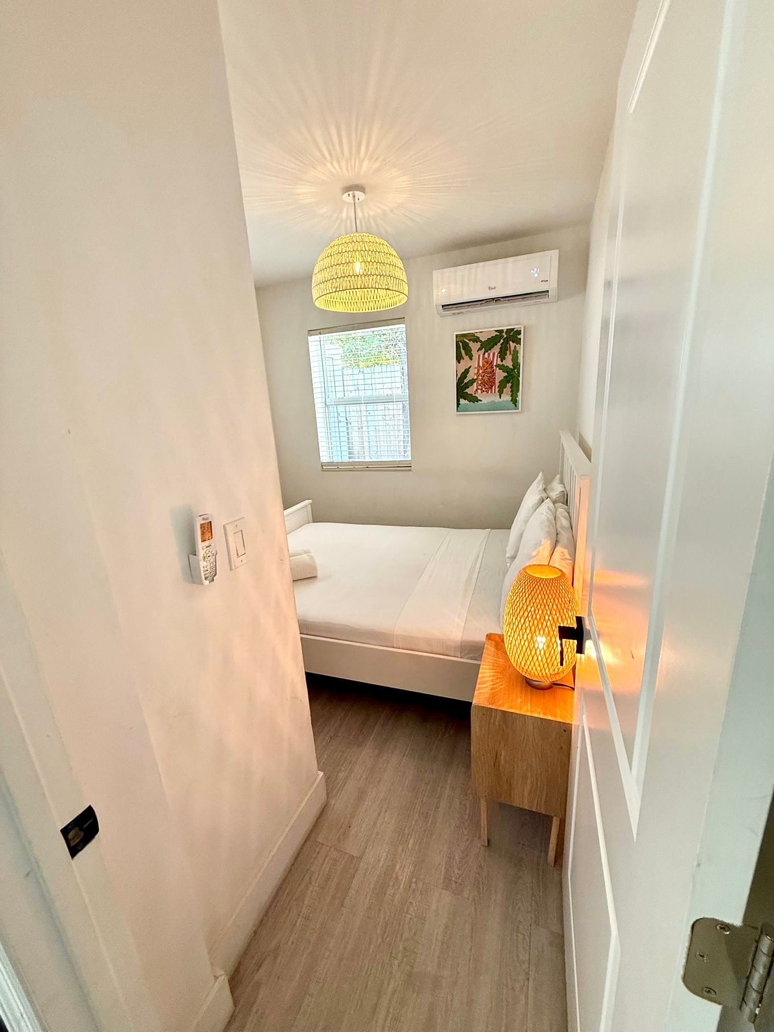 Narrow bedroom with white walls, bed, nightstand, lamp, and hanging light.