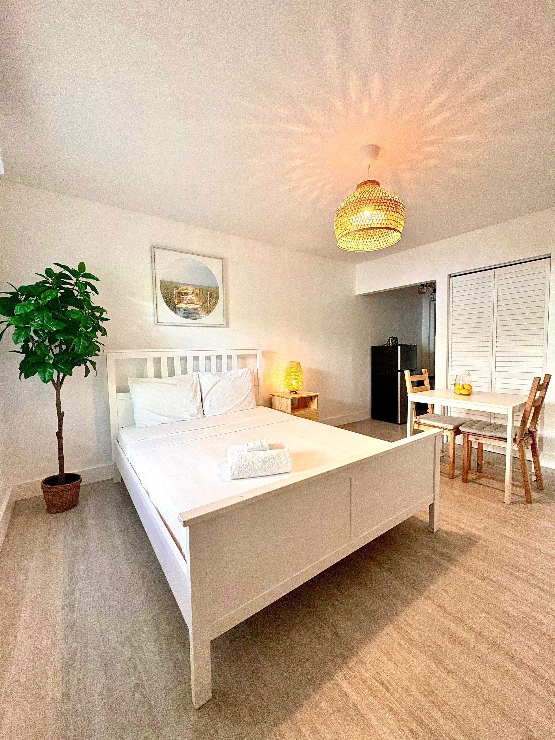 White-walled bedroom with white bed, art, lamp, and table. A potted plant is to the left.