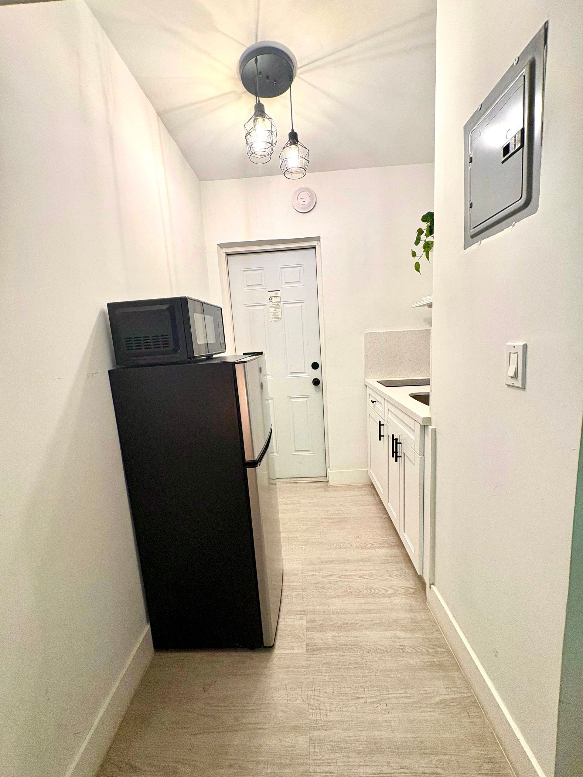 Narrow kitchen with refrigerator, microwave, white cabinets, door, and hanging lights.