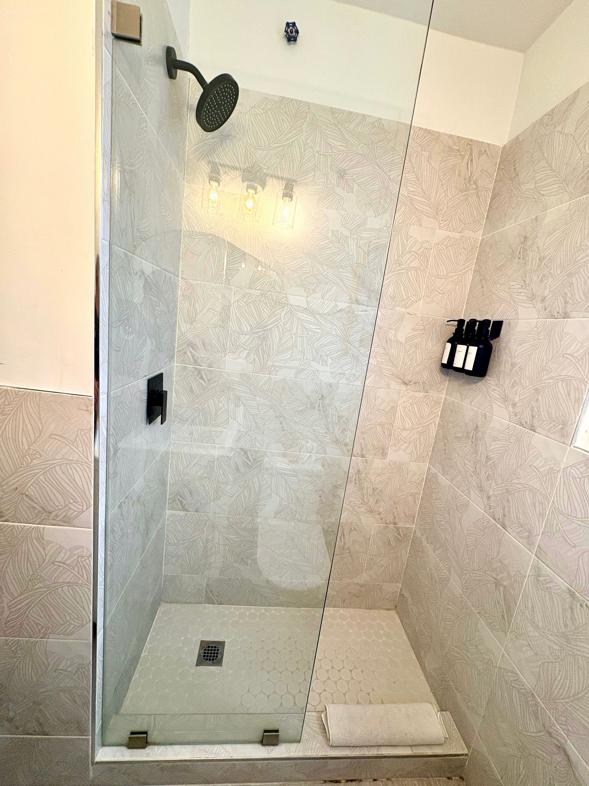 Shower stall with glass door, black fixtures, and light gray tile walls. Towel on a bench.