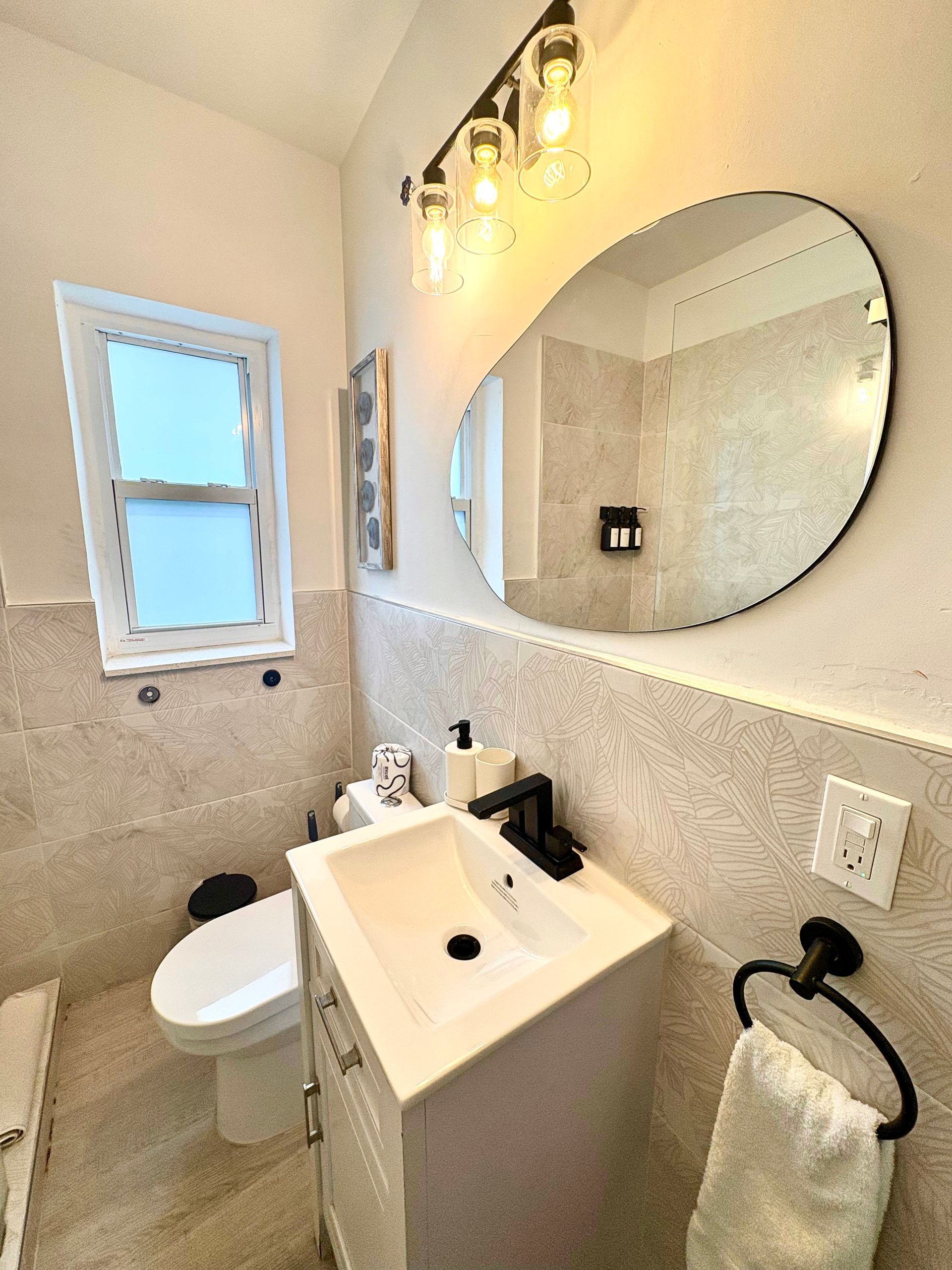 Bathroom with off-white textured tile, white sink, black fixtures, oval mirror, window, toilet.