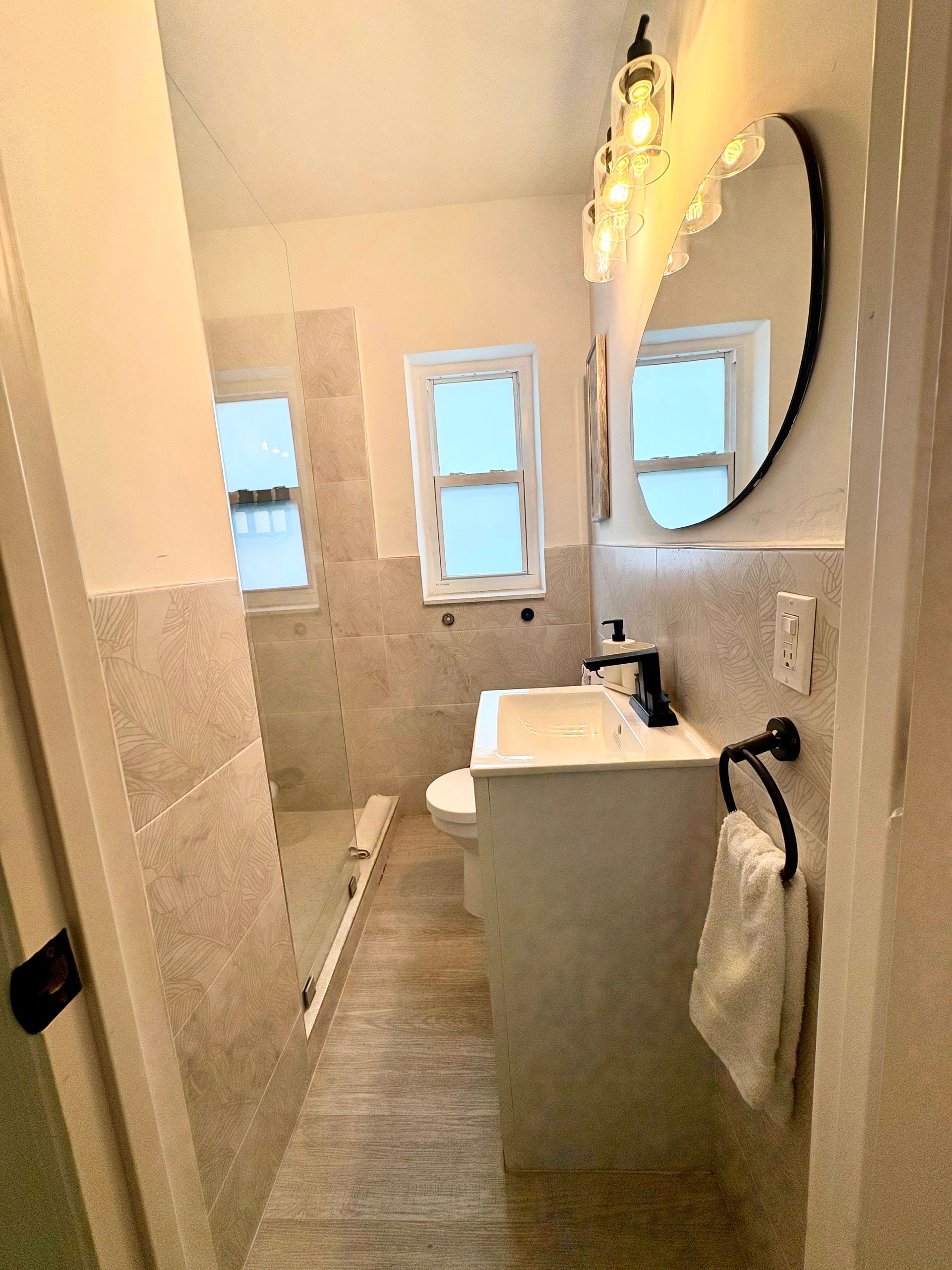 Small bathroom with white vanity, black faucet, oval mirror, and light-colored tile.