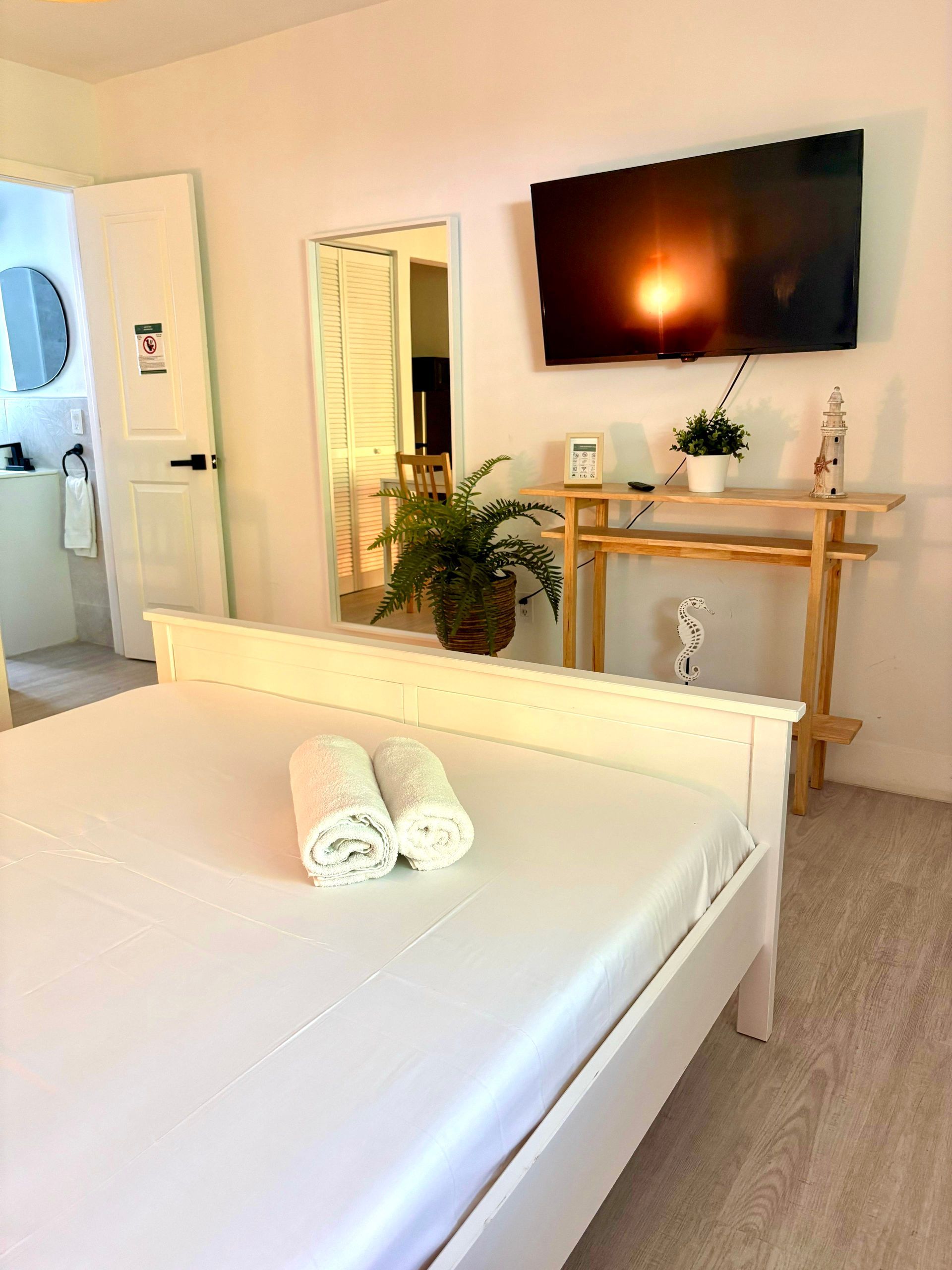 White bed in a bright room with a TV, mirror, and small table with decor.