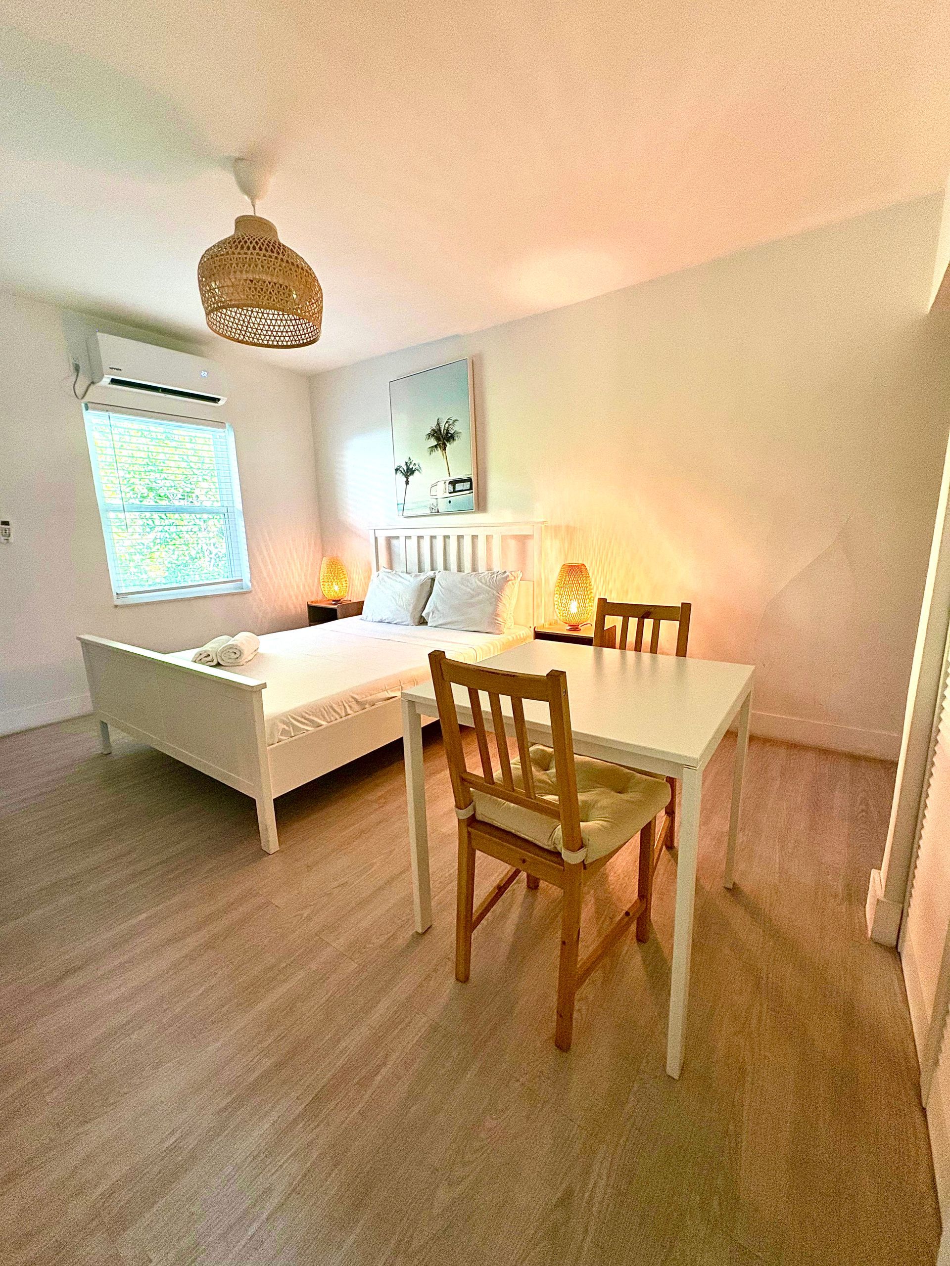 Bedroom with white bed, table, chairs, woven lampshade, and window.