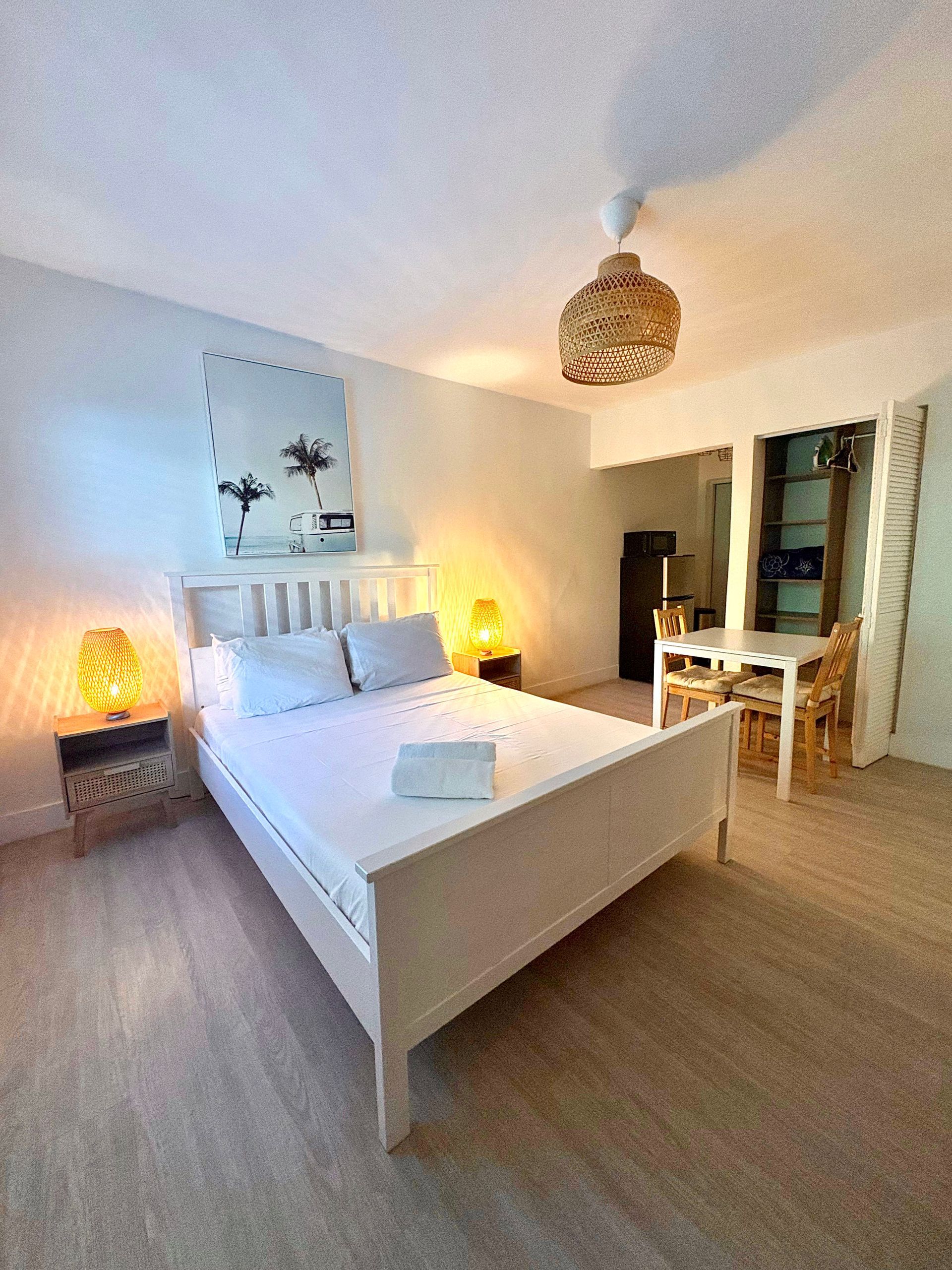 Bedroom with white bed, artwork, bedside lamps, and woven lampshade. Dining area visible.