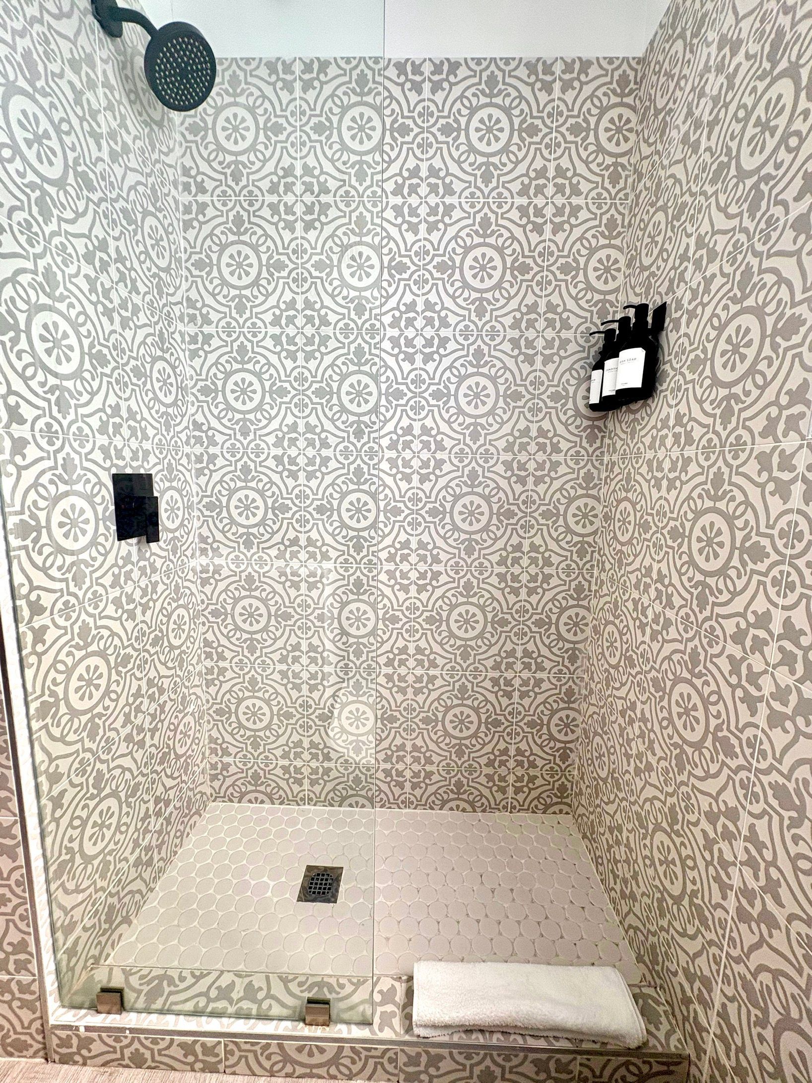 Shower with gray patterned tile walls, glass door, and a white towel on the floor.