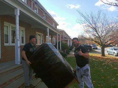 Two men are carrying a large black object in front of a brick building.