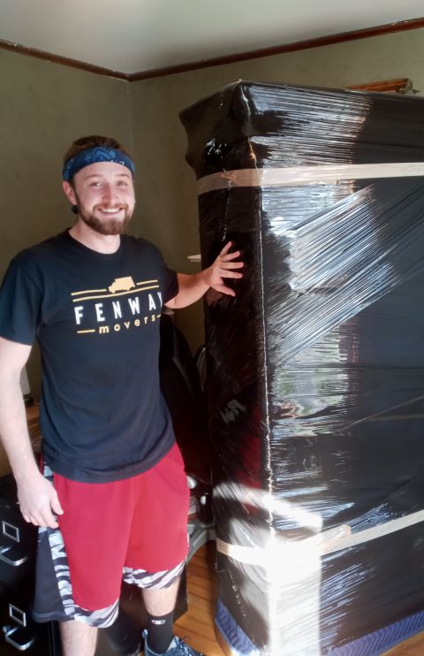 A man in a black shirt is standing next to a black box wrapped in plastic.