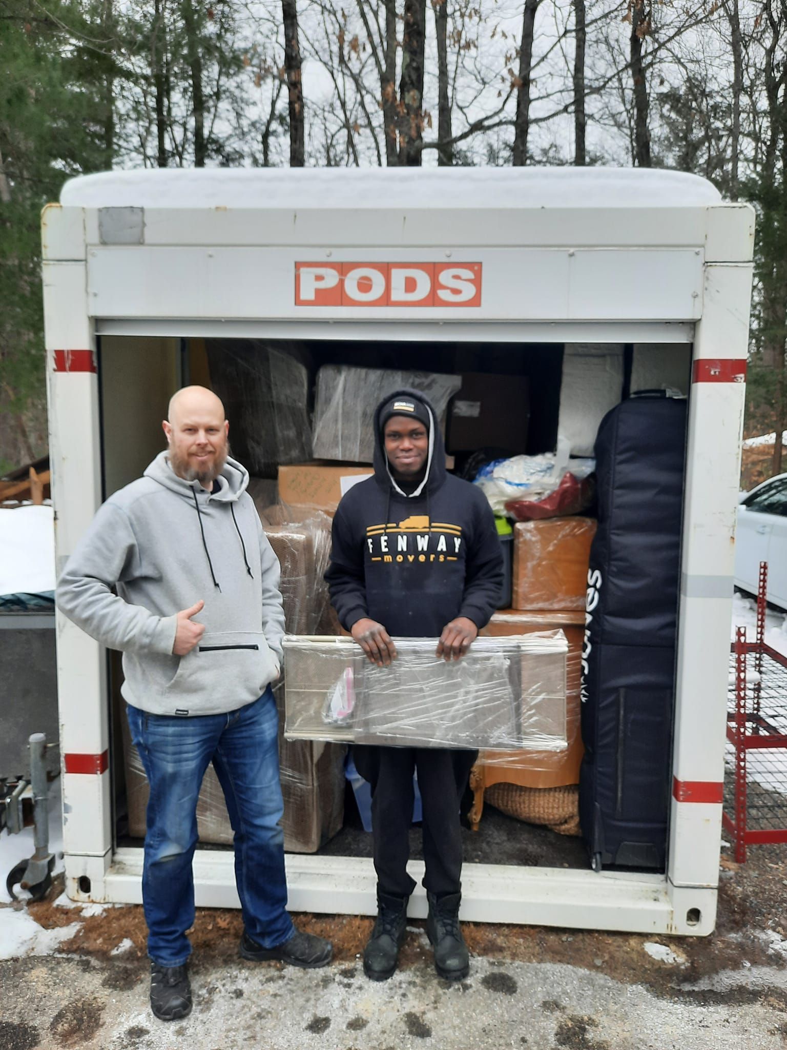 Two men are standing in front of a pods trailer holding boxes.