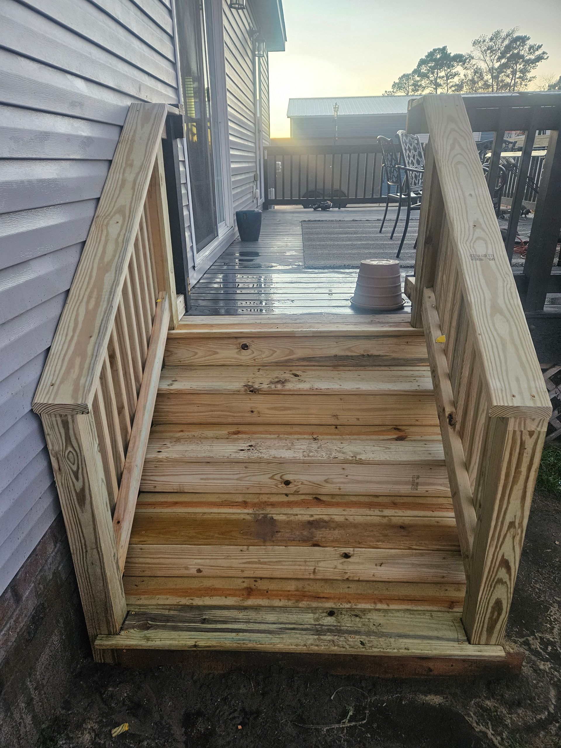 Wooden outdoor steps with railings leading up to a deck, near a house with gray siding.