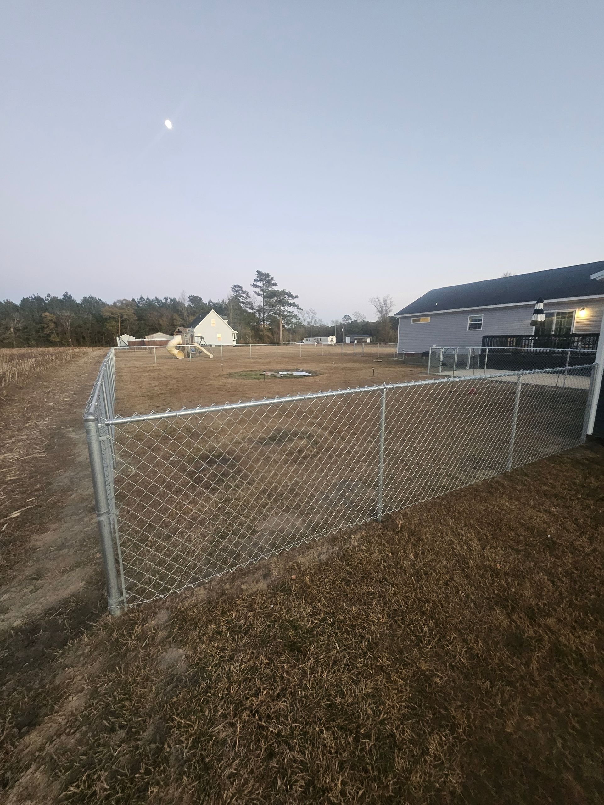 Chain-link fence surrounding a grassy yard with houses in the background under a dusky sky and moon.