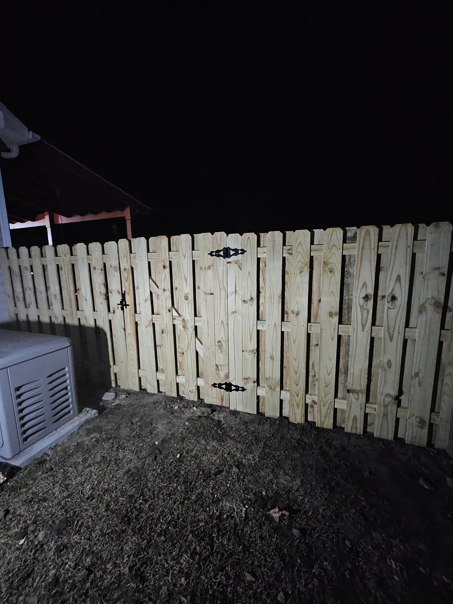 Wooden fence with gate, lit at night, next to an AC unit and a dark yard.