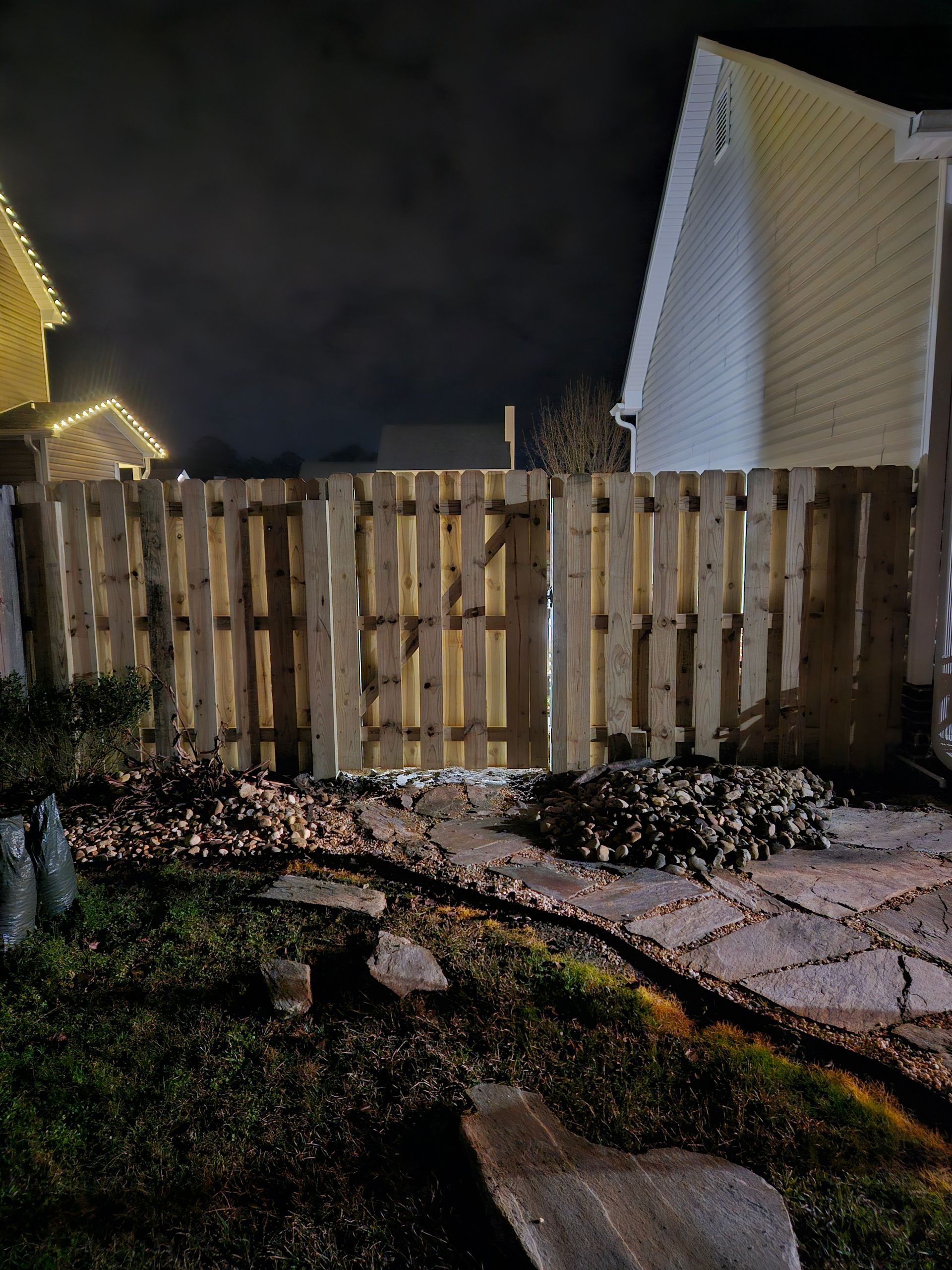 Wooden fence at night, lit by unseen light source, in front of houses with dark sky overhead.