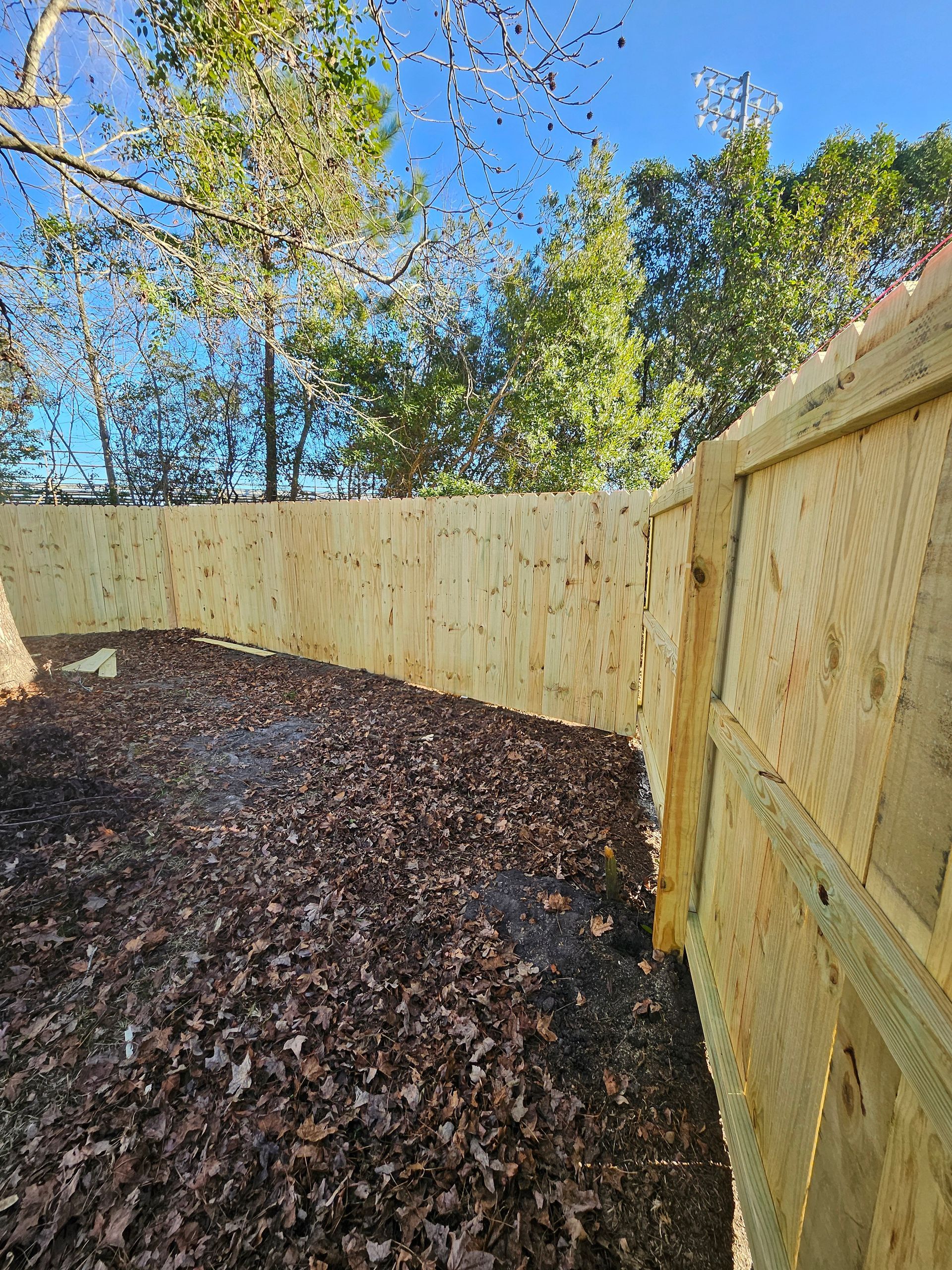 Wooden fence surrounding a yard with fallen leaves. Trees and blue sky visible in the background.