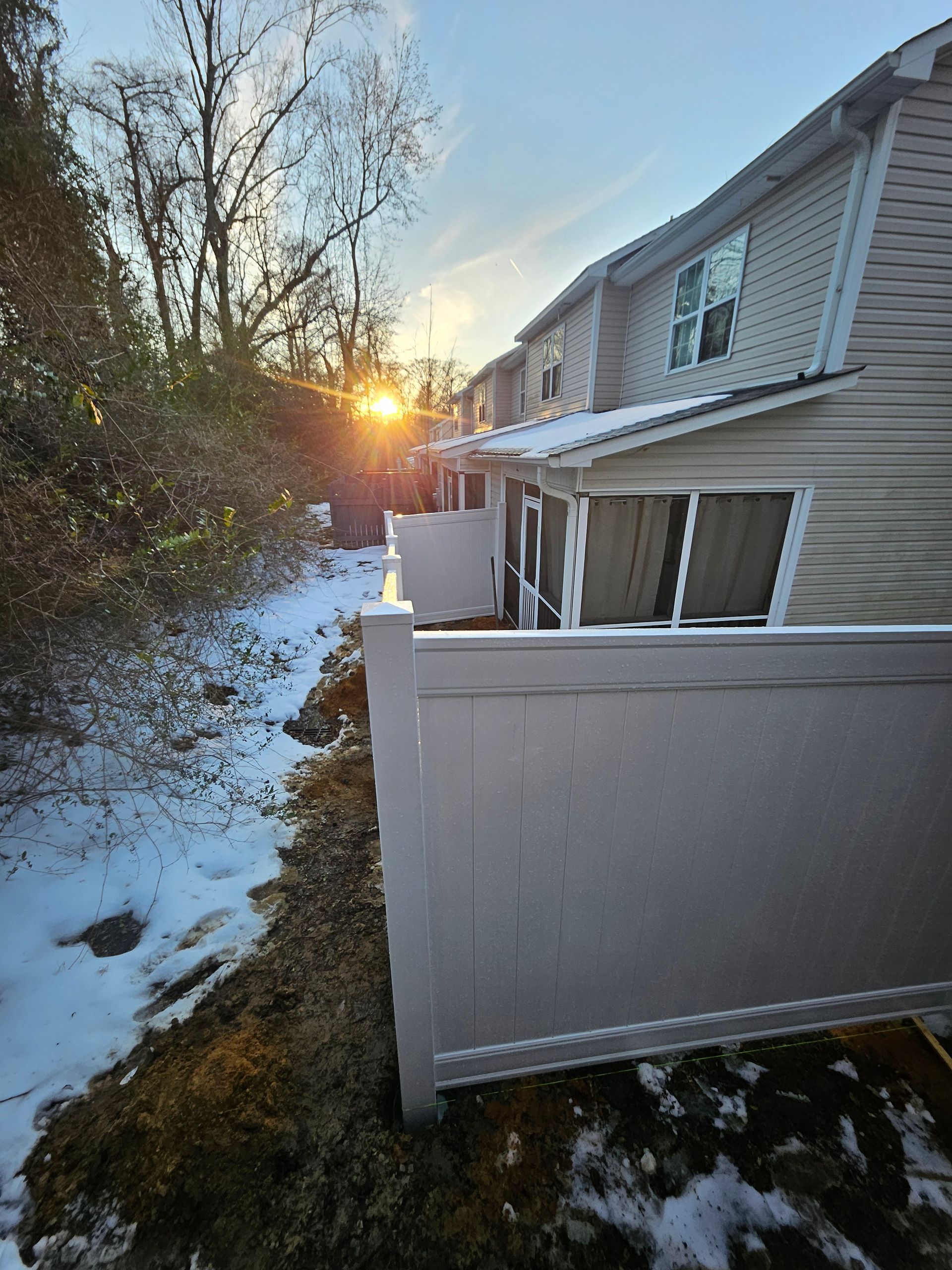 Snowy townhouse exterior with a fence. Sun setting in the distance.