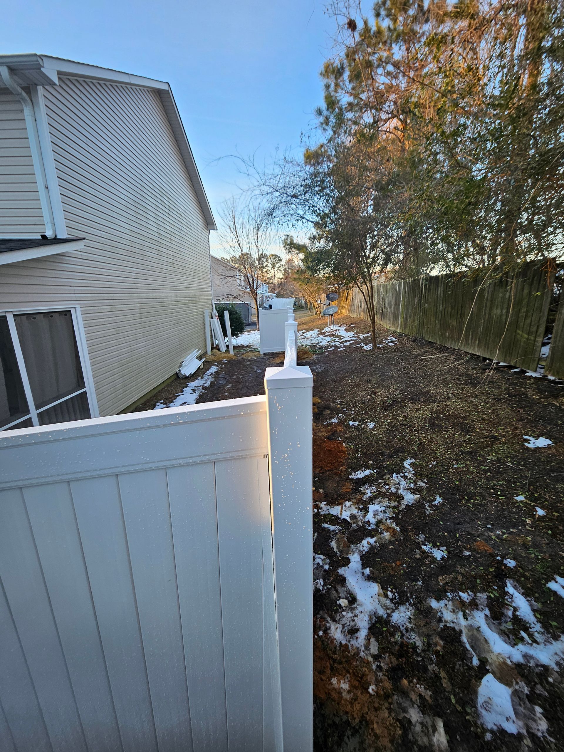 Snowy backyard scene with white fence, buildings, and a tree under a bright blue sky.
