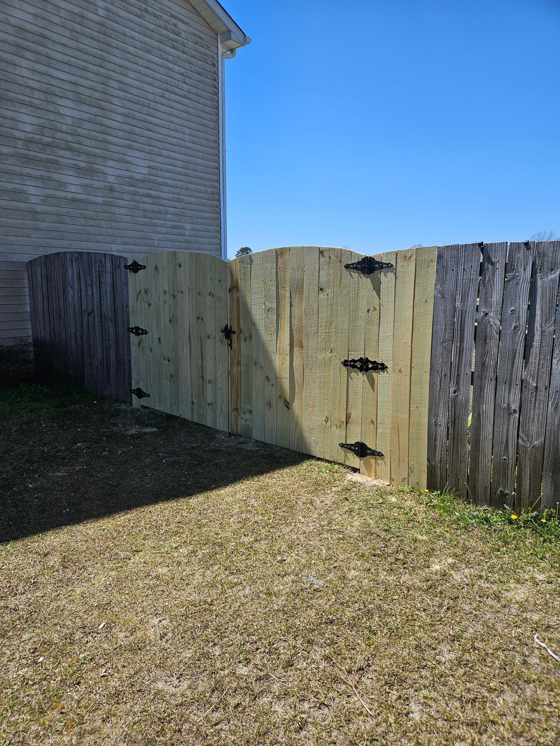 Wooden fence gate with black hinges, next to a brick building.