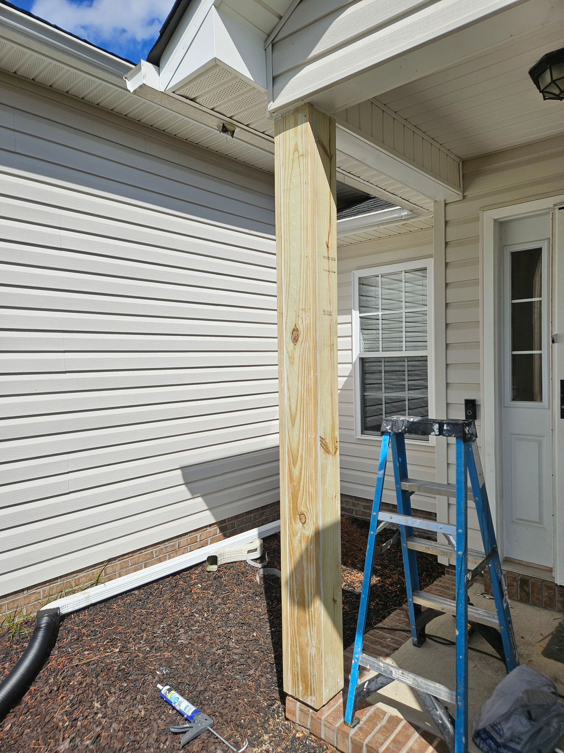 A wooden post on a porch, siding wall. A blue ladder and door are visible. Brown mulch and a gutter system are also present.