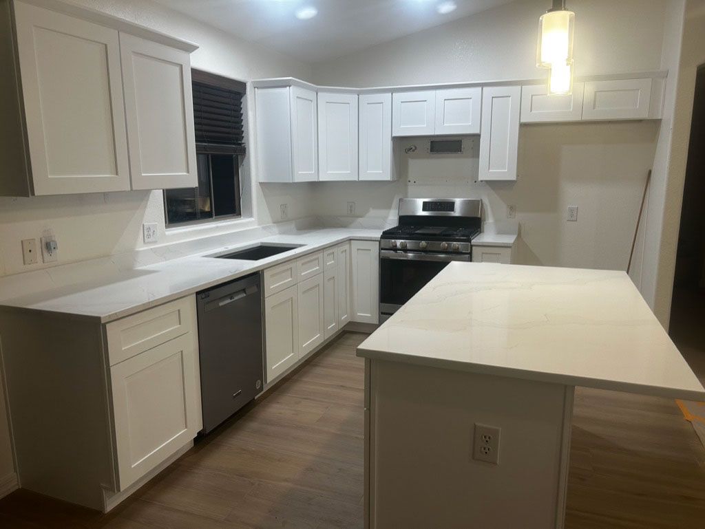 White kitchen with a stove, island, cabinets, and countertops. The floor is wood, and there's a window with dark blinds.