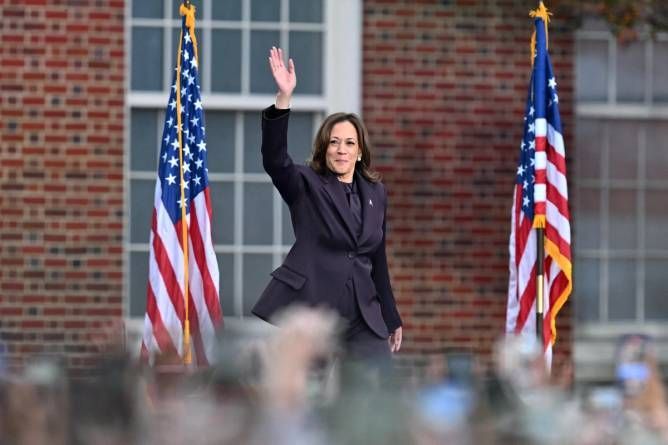 A woman in a suit is waving at a crowd in front of american flags.