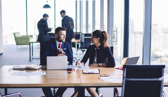 A man and a woman are sitting at a table in an office talking to each other.