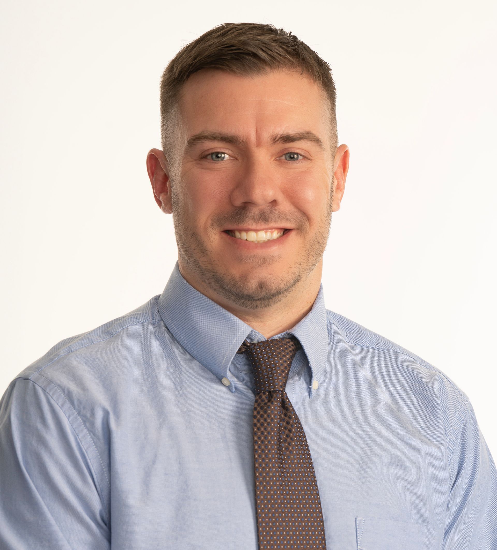 A man in a blue shirt and brown tie is smiling for the camera.