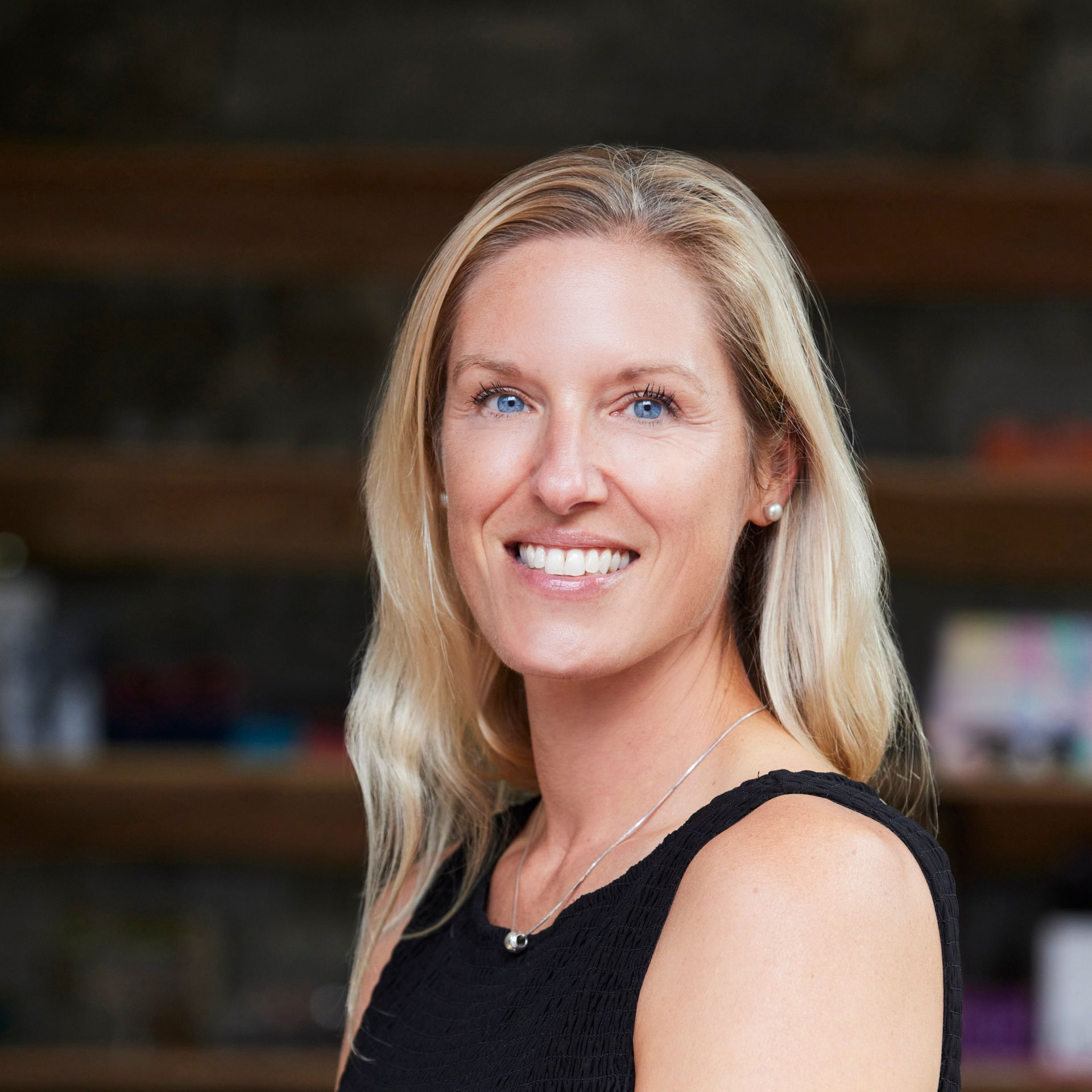 Blonde woman smiles at camera, wearing black dress, in front of a bookshelf.