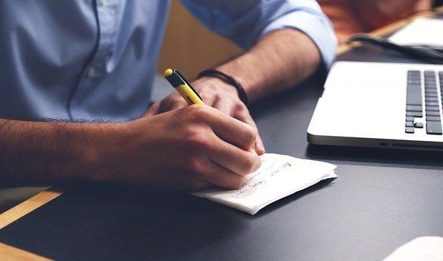 A man is writing on a notepad next to a laptop.