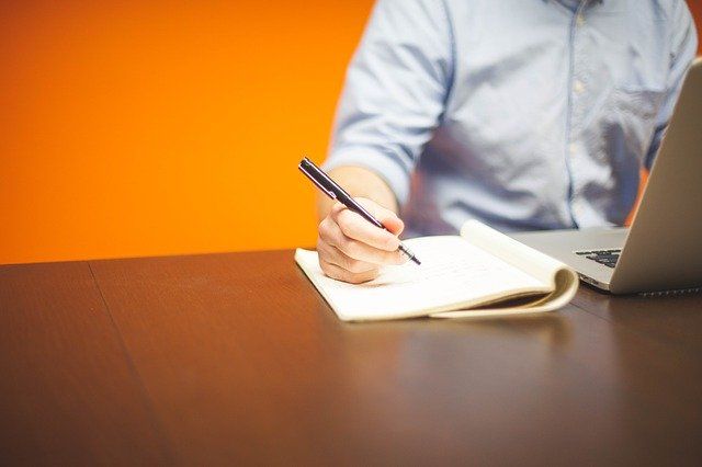 A man is sitting at a desk with a laptop and writing in a notebook.