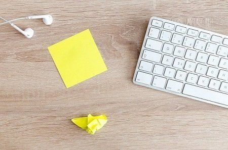 A keyboard , headphones , a yellow sticky note , and a piece of paper on a wooden table.