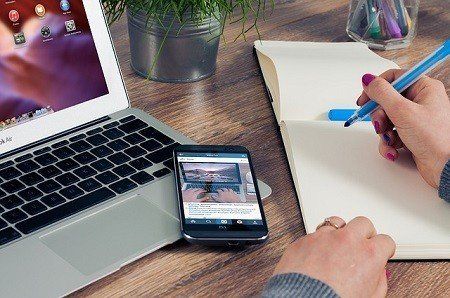 A person is writing in a notebook next to a laptop and a cell phone.