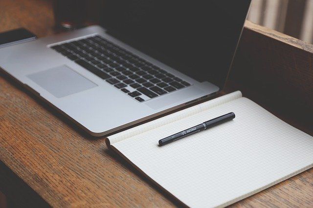 A laptop is sitting on a wooden desk next to a notebook and pen.