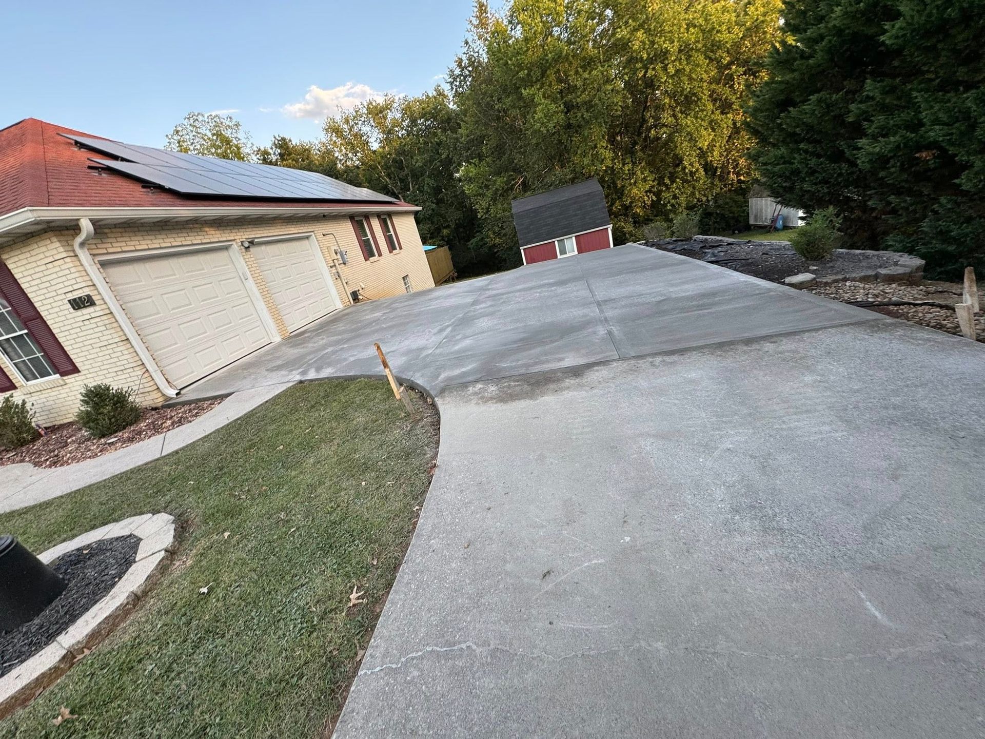 Driveway slopes up towards a house with solar panels, green grass, and trees under a blue sky.