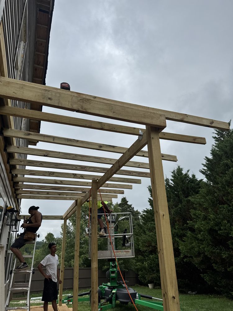 Workers constructing a wooden pergola on the side of a house, using ladders and lifts outdoors.
