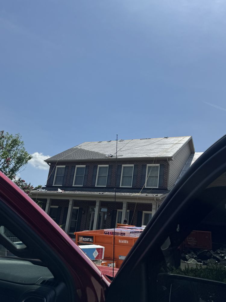 A two-story house with roofing work in progress, viewed from inside a red car. Bright blue sky.