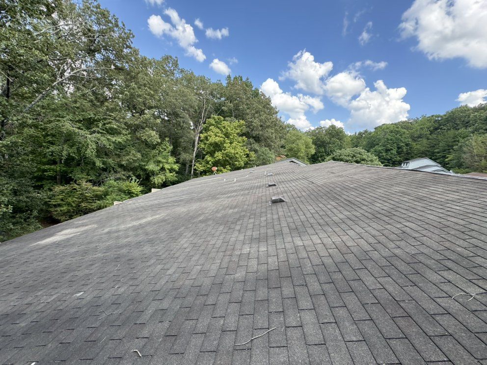 Grey asphalt shingle roof, with trees in the background, under a partly cloudy blue sky.