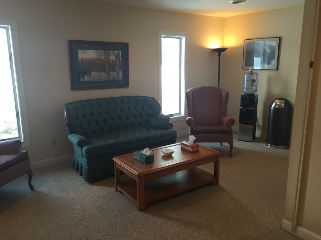 Waiting room with teal sofa, armchair, coffee table, art, water cooler, and trash can.