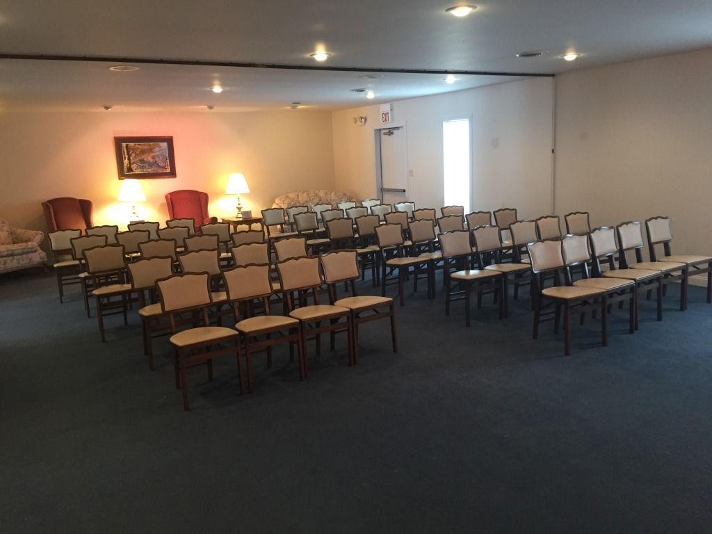 Rows of chairs face forward in a large room. The room has neutral walls, carpet, and an exit door.